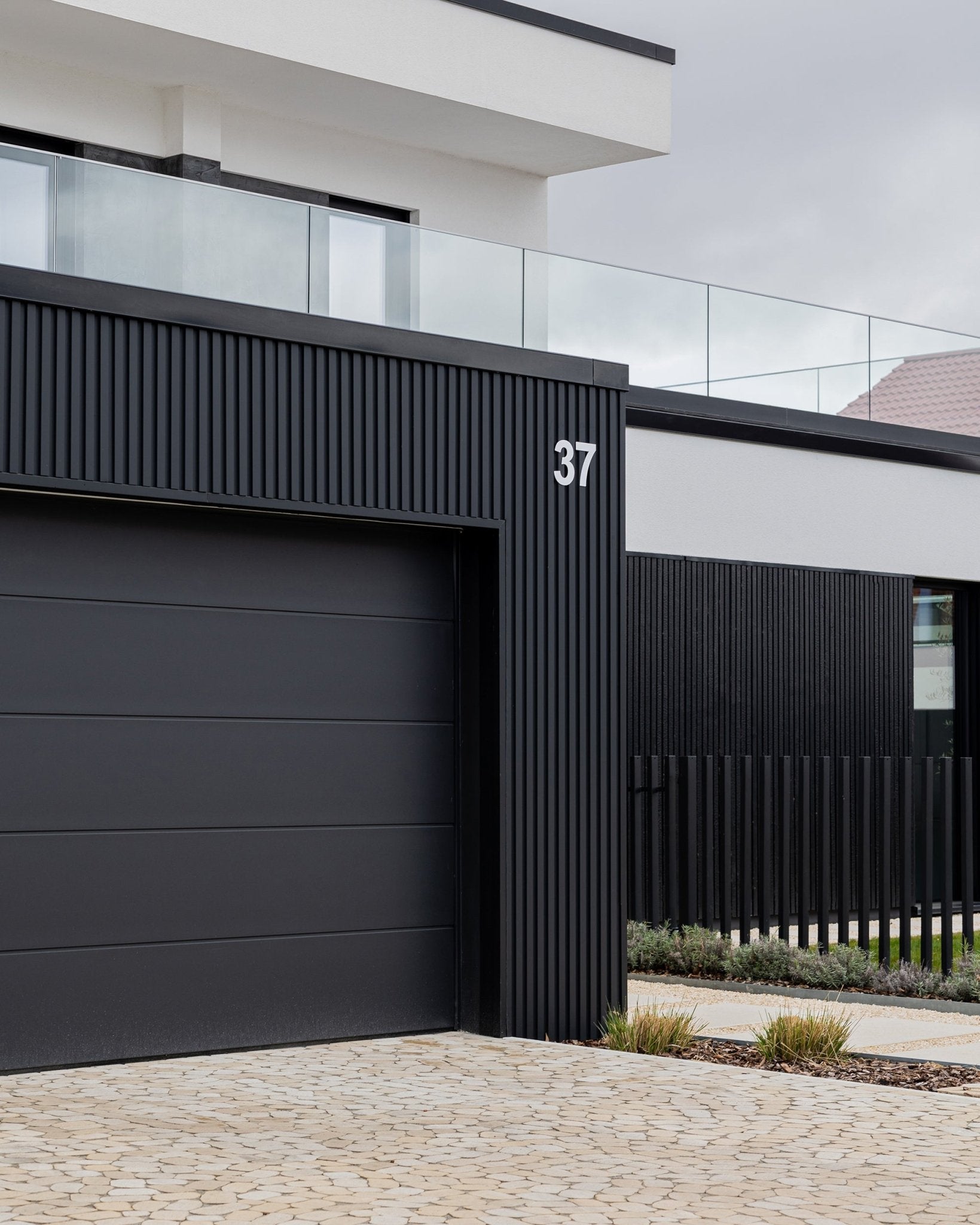 Black och vitt husfasad med WoodUpp Aluwood fasadpaneler, black garageport, vertical ribbdetalj, husnummer 37 och balkongräcke i glas.